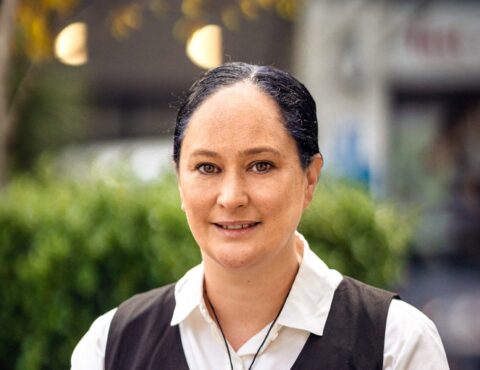 A woman with black hair tied back stands outdoors, smiling confidently. She wears a white collared shirt layered under a black vest. The background has soft, green foliage, giving a professional yet relaxed appearance.