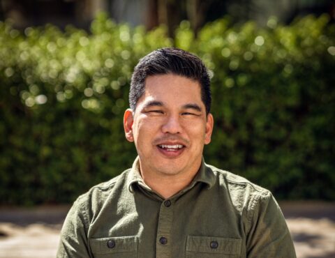 A smiling man wearing a green button-up shirt sits outdoors. His short black hair is neatly styled, and the background features blurred green foliage, suggesting a natural, sunny environment.