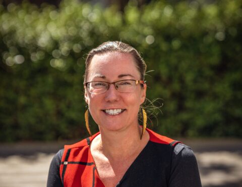 A close-up outdoor portrait of a woman wearing glasses and golden feather earrings. She has light brown hair tied back and is smiling confidently. She wears a black top with red accents, and the background is a softly blurred green hedge.