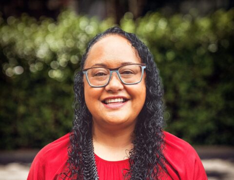 A cheerful portrait of a woman with curly black hair and glasses, wearing a bright red top. She smiles warmly, and the blurred background consists of green foliage, adding vibrancy to the image.