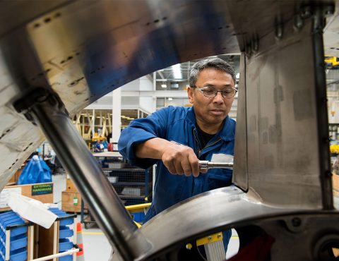 A male technician wearing safety glasses and a blue work uniform uses a tool to repair or assemble a large piece of industrial machinery. He works in a spacious workshop with visible tools, parts, and equipment in the background.