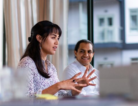 A woman with long dark hair gestures as she speaks during a discussion. She is wearing a white blouse with black polka dots. A smiling man with a white shirt listens attentively in the background, with large windows showing a modern building exterior.