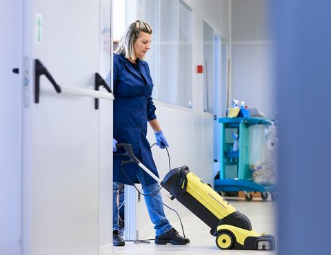A cleaning professional wearing a navy-blue uniform and gloves operates a yellow floor cleaning machine in a brightly lit hallway. Cleaning supplies on a trolley are visible in the background, indicating a professional cleaning environment.