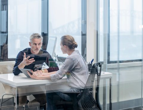 Two people sitting in an office. There is a large glass door in front of them.