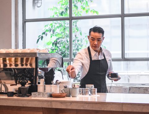 A person making coffee. They are wearing a barista uniform. In the background it a large window.