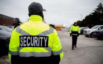 A person with their back away from the camera. The back of their top reads security. In the background is a person walking towards then and a carpark.