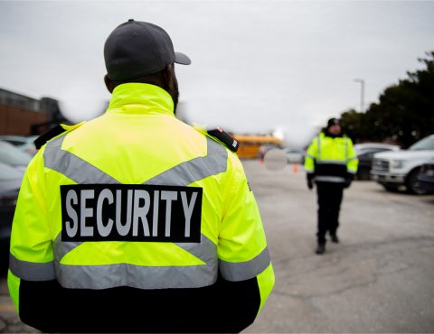 A person with their back away from the camera. The back of their top reads security. In the background is a person walking towards then and a carpark.