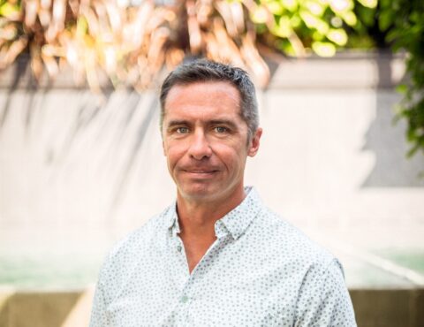 A portrait of a middle-aged man outdoors. He has short salt-and-pepper hair and a subtle smile. He wears a light-colored, patterned button-up shirt. The background is softly blurred with sunlit foliage.