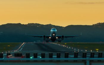 A striking image of a large airplane taking off at sunset. The plane is centered on the runway, surrounded by lights, with silhouetted hills in the background and an orange sky.