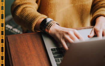 A close-up photo of a person typing on a laptop. The person wears a mustard-colored sweater and multiple bangles on their wrist. To the left of the image, there are Māori-inspired orange and black designs, adding cultural detail.