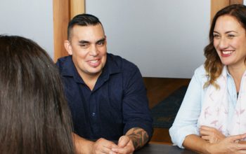 Three people sitting down and having a meeting. They are dressed in semi formal attire.