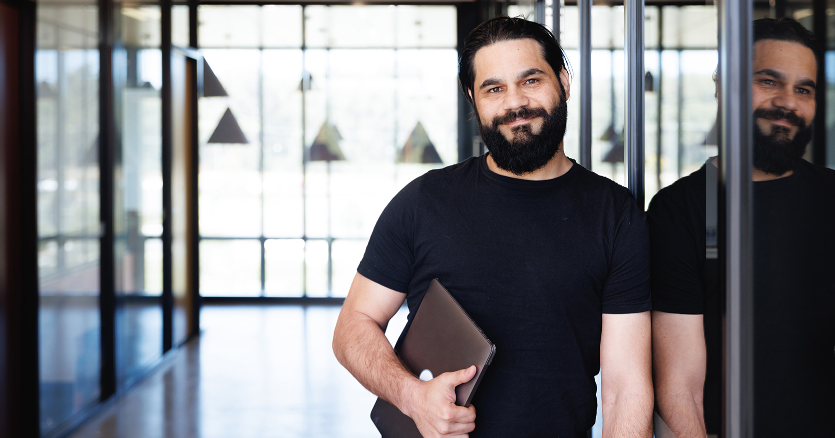 A person leaning on a wall with their image reflected on the glade next to them. In the background is empty hall way with big windows.