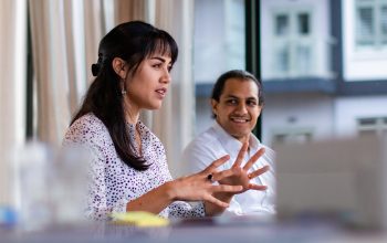 Two people sitting down. One is explaining something with their hands while the other person looks at them.