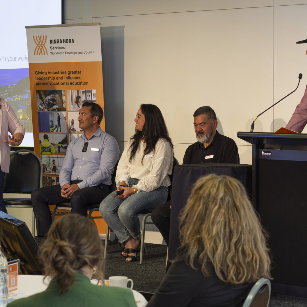 A group discussion panel featuring four people sitting on chairs on stage. A woman in a white blazer stands and gestures while speaking to a man in a pink blazer and black hat, who is behind a podium with a microphone. Audience members are visible in the foreground.