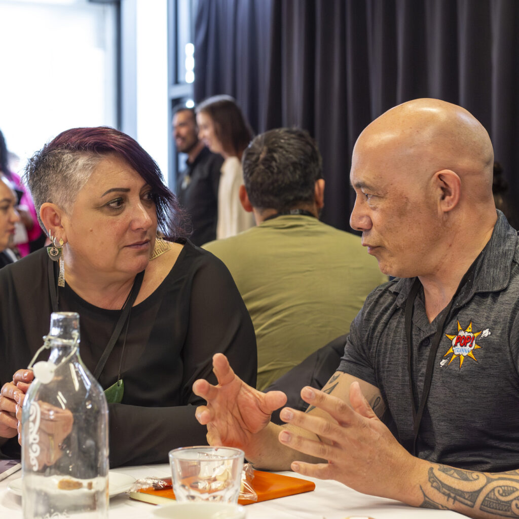 A woman with a shaved hairstyle dyed purple listens intently to a bald, tattooed man wearing a black shirt with a logo. Both are seated at a table with water bottles and glasses.