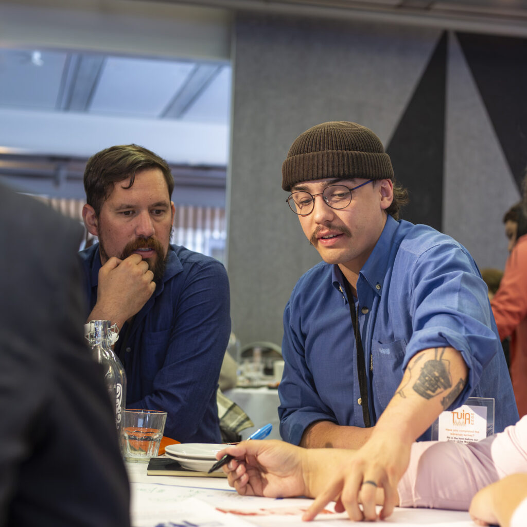 A group of people sitting at a table, collaborating. A man in a blue beanie and glasses points at a document while speaking, with others, including a man in a blue shirt and a woman with glasses, looking on.