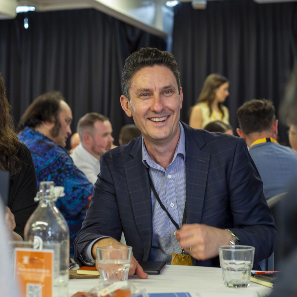 A man in a navy suit smiles while seated at a table. Other attendees are visible in the background, engaged in conversation, with dark curtains in the room.