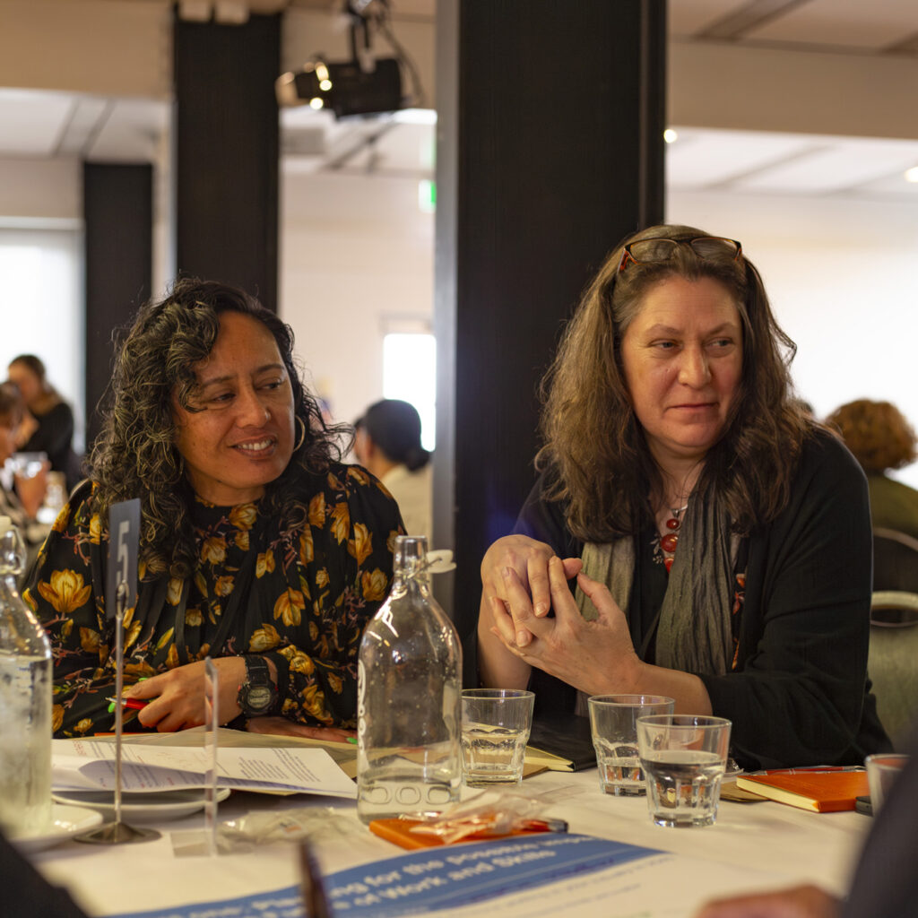Two women sitting at a table having a conversation. One woman, wearing a floral dress, smiles while the other woman, in black, leans slightly forward. Bottles of water and notes are on the table.