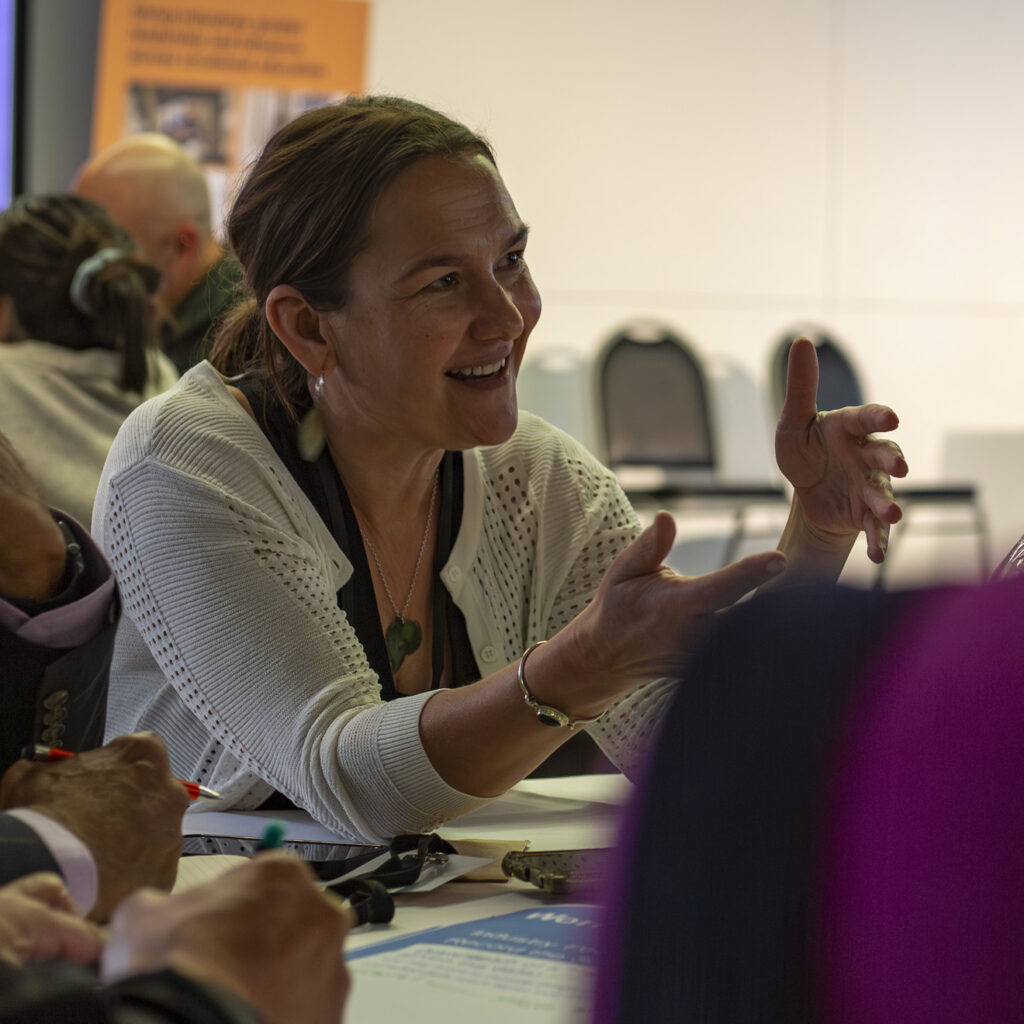 A woman in a white cardigan sits at a table, smiling and animatedly gesturing with both hands as she speaks. Other people and water bottles are visible in the background.
