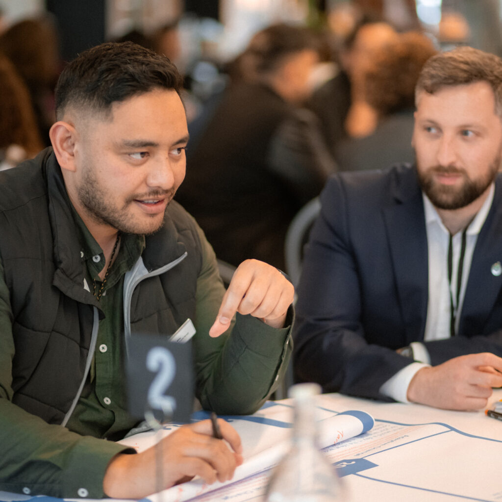 Two men are sitting at a table having a discussion. One man, wearing a dark green shirt and black vest, is speaking and gesturing, while the other man in a navy suit listens attentively. A table number "2" is placed in front of them.