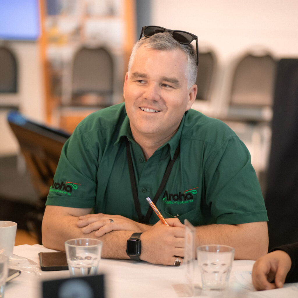 A man in a green "Aroha Helicopters" shirt is sitting at a table smiling and holding a pen. Glasses of water and coffee mugs are on the table.
