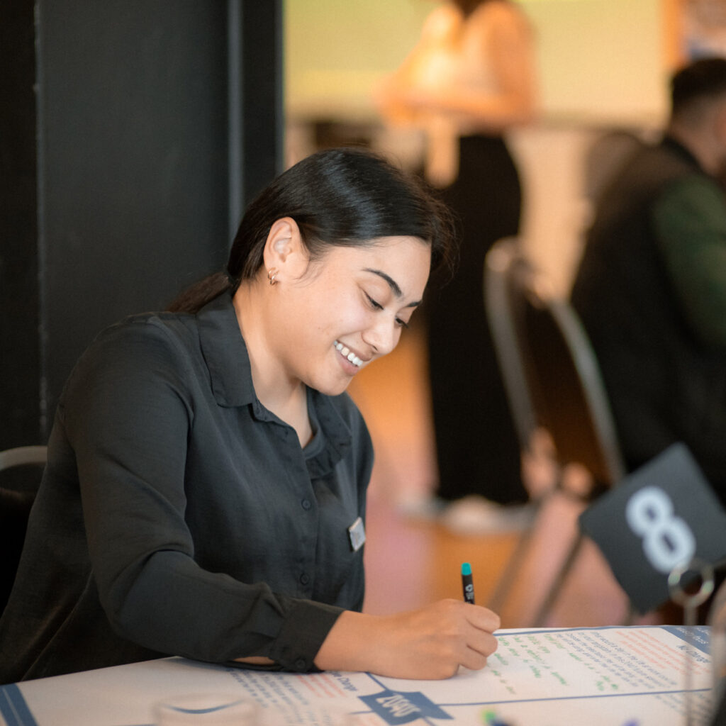A woman with dark hair in a black shirt is sitting at a table, smiling and writing on a large sheet of paper. A small table number "8" sign is visible in front of her.