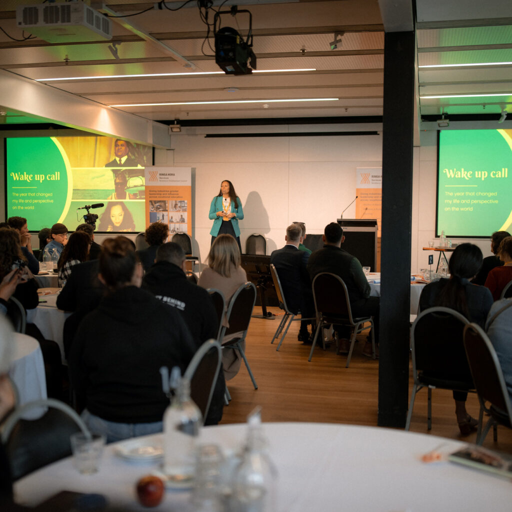 A wide view of a conference room with attendees sitting at tables listening to a speaker on stage. The speaker, a woman in a teal blazer, is presenting with two large screens displaying the title “Wake up call” and visuals.