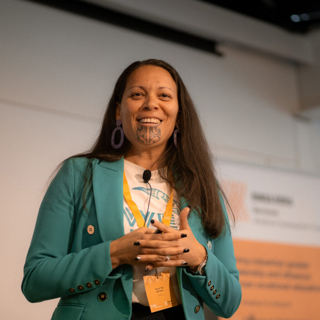 A close-up of the woman in the teal blazer standing on stage and smiling. She has long hair, purple earrings, and a traditional Māori tā moko chin tattoo. Image 9: A man in a light blue suit is speaking at a podium with a microphone, smiling and gesturing with both hands. A Ringa Hora Workforce Development Council banner is behind him.