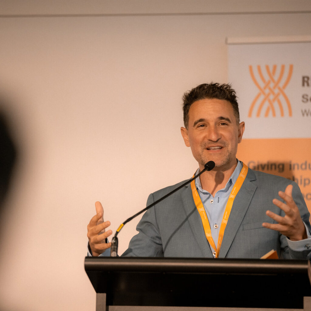 A man in a light blue suit is speaking at a podium with a microphone, smiling and gesturing with both hands. A Ringa Hora Workforce Development Council banner is behind him.