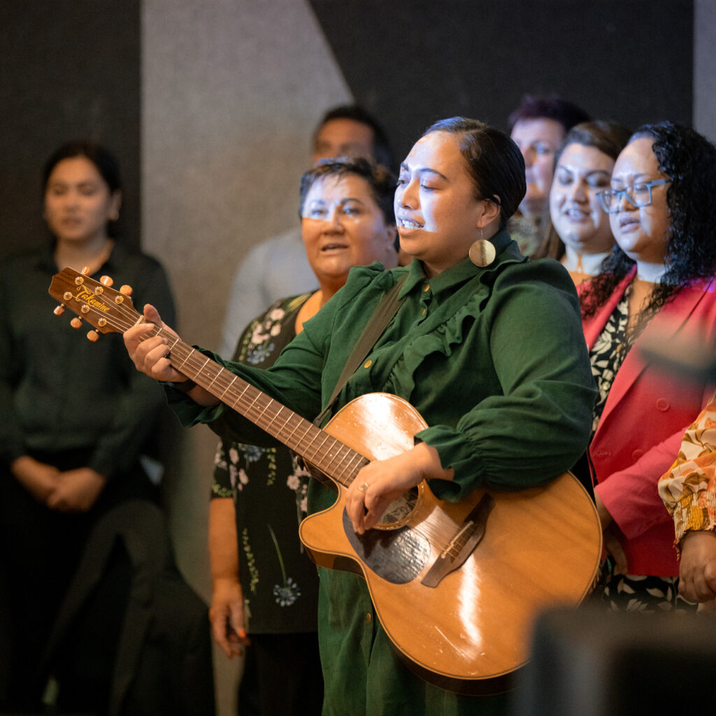 A group of people stand together singing, led by a woman in a dark green outfit playing a wooden acoustic guitar. The group includes women wearing floral and bright-colored clothing, singing passionately.