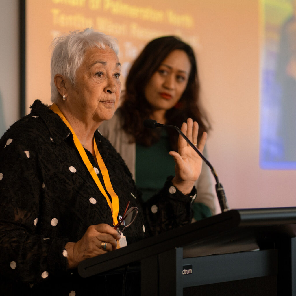 The same white-haired woman from the previous image stands at the podium with another woman in a green outfit slightly behind her. She gestures while speaking, with an orange presentation screen in the background.