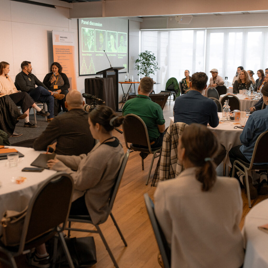 A wide view of a panel discussion in a conference room. Five people are seated on stage, speaking to an audience seated at round tables. A screen displaying "Panel discussion" with participant names and photos is visible.