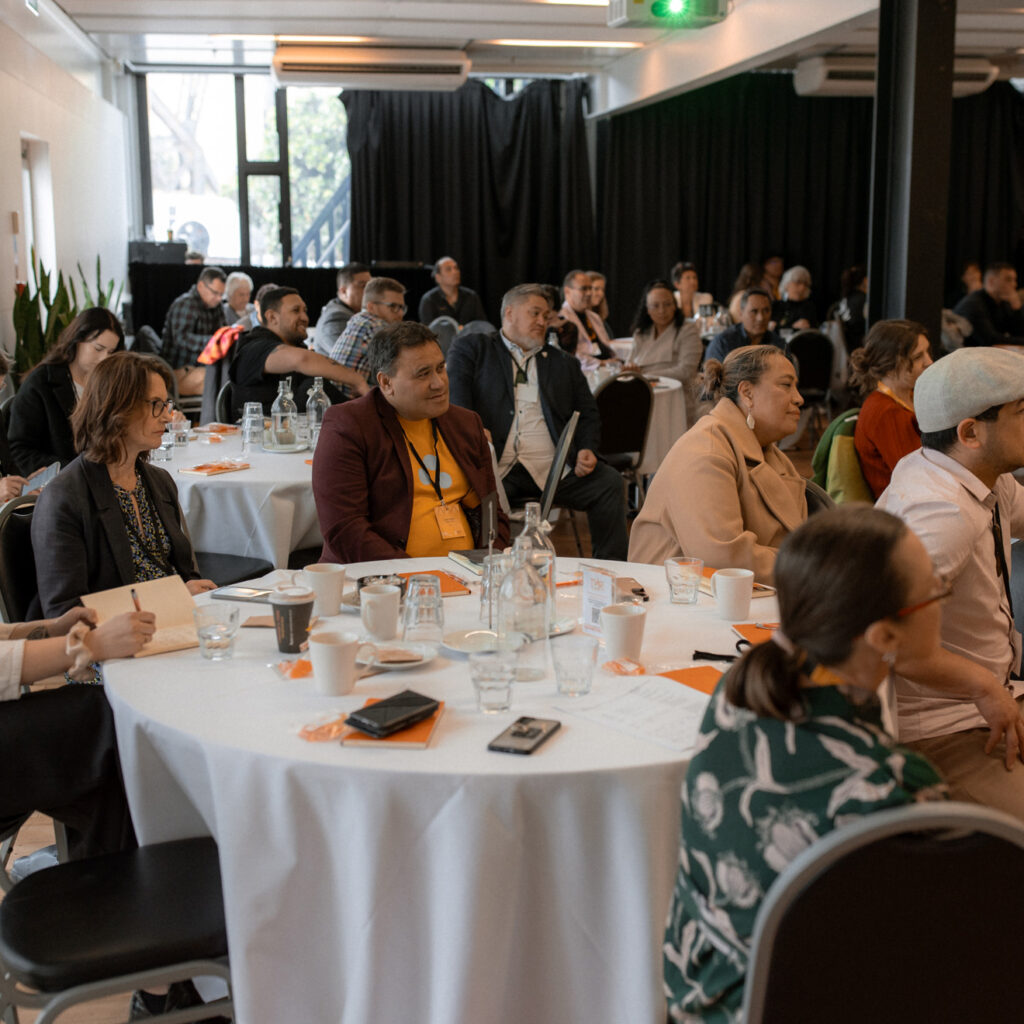 A diverse group of attendees seated at round tables in a conference room, listening intently. Papers, drinks, and orange name tags are scattered across the tables.