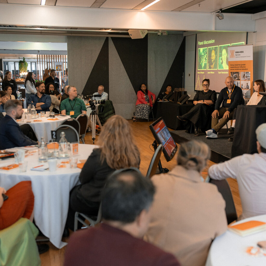 A wide-angle view of a panel discussion in progress, with panelists seated on stage and an engaged audience. A presentation screen behind the panelists displays "Panel Discussion" with speaker images.