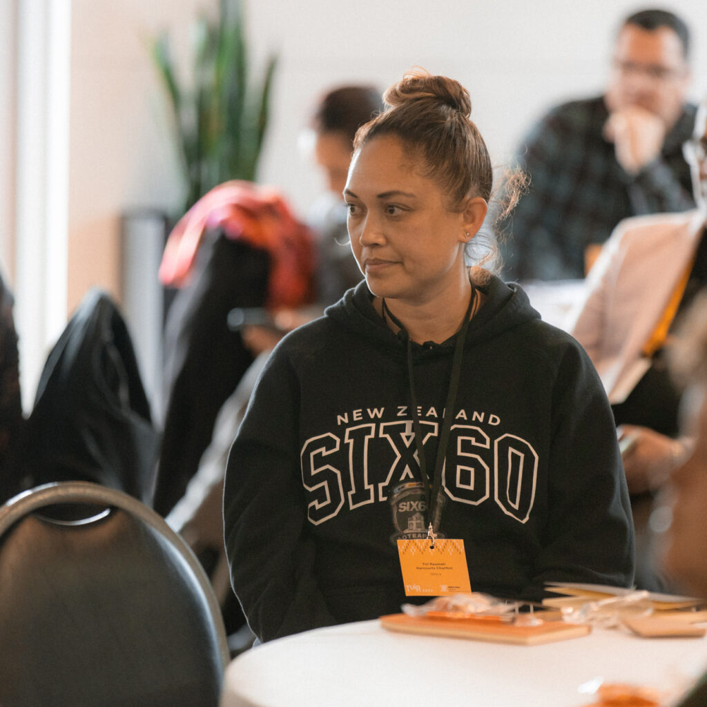 A woman in a "New Zealand SIX60" hoodie sits at a round table, appearing attentive during the event. Other attendees are visible in the background.