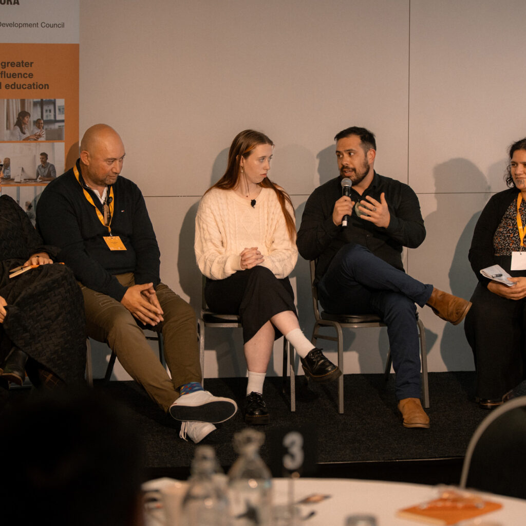 A group of five panelists sits in a row on stage during a panel discussion. One speaker holds a microphone while others listen attentively.