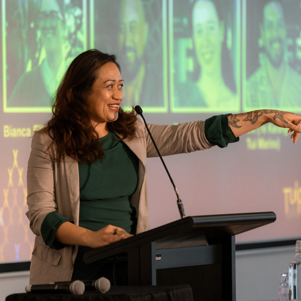A woman in a green dress points energetically from a podium while speaking. The background screen shows blurred images of people.