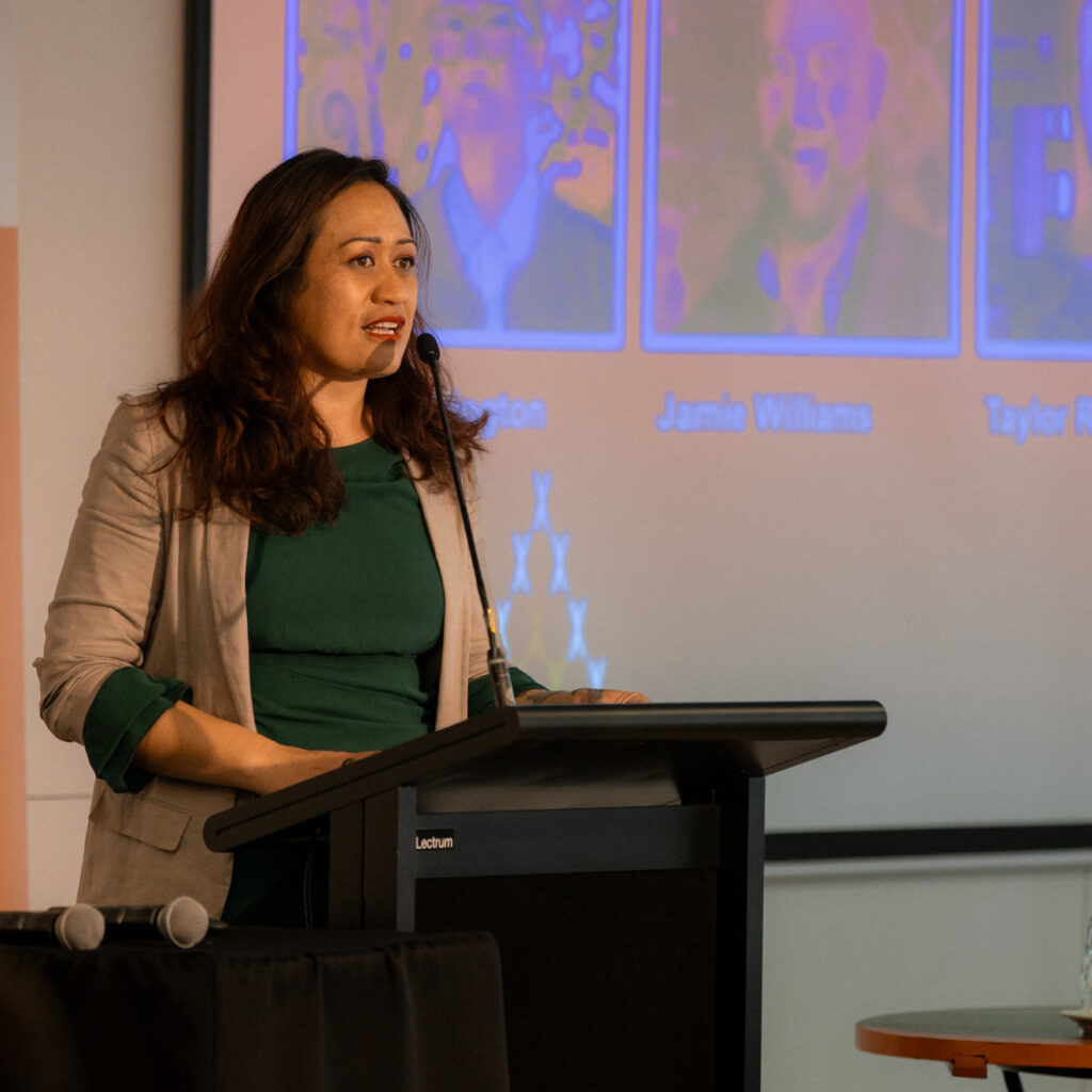 A woman at a podium speaks confidently to the audience. Behind her, a presentation screen displays speaker information.
