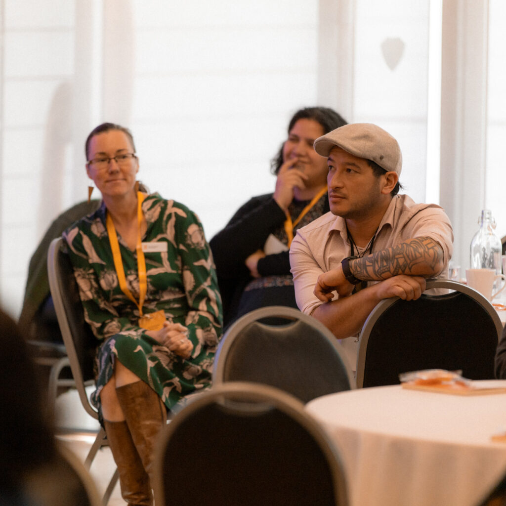 Two attendees, a woman in a green floral dress and a man in a flat cap with visible arm tattoos, sit at a table watching a presentation.