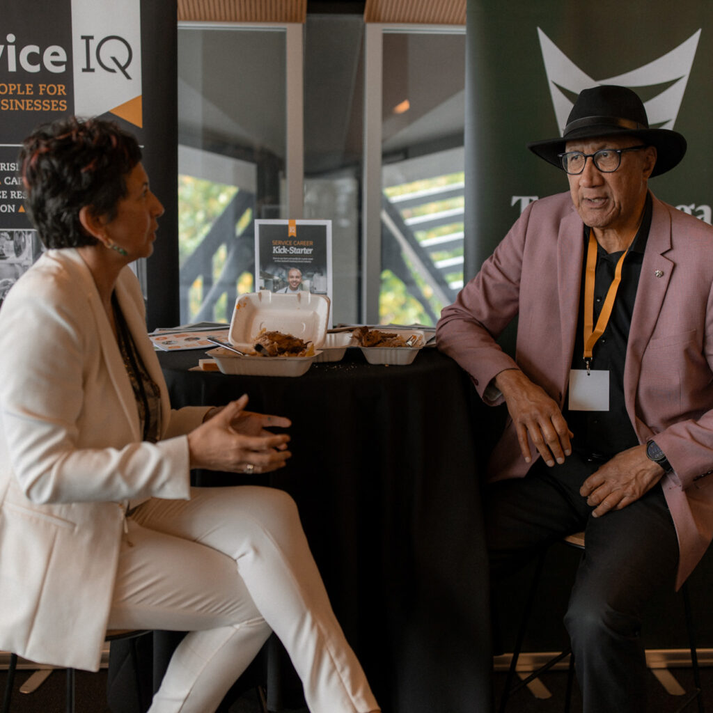 Two individuals sit at a table for a discussion near Service IQ and Te Ara Hora banners. Food containers and promotional materials are visible on the table.