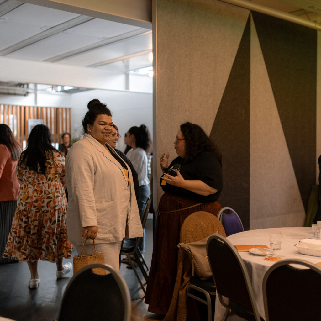 Attendees are gathered near tables and standing areas, conversing and networking. A woman in a beige suit smiles while holding a woven bag.