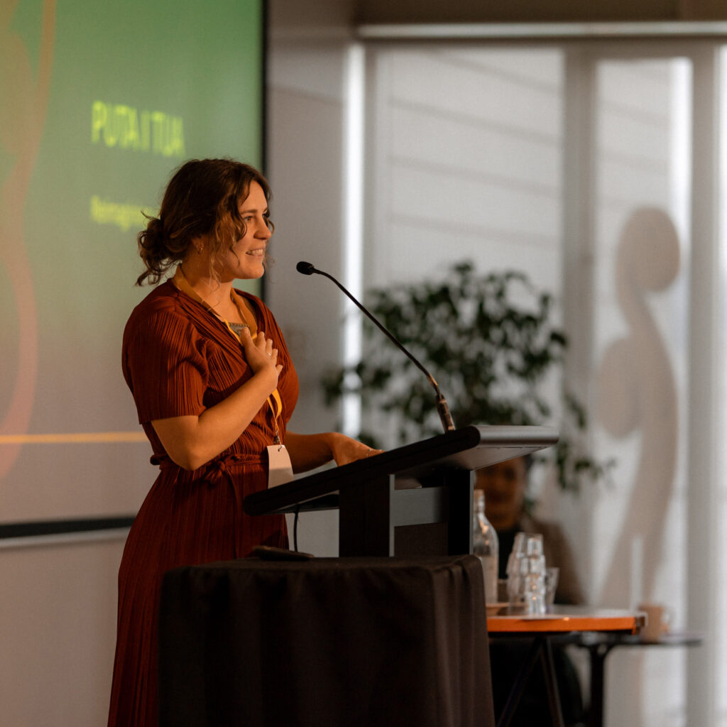 A young woman with curly brown hair wearing a rust-red dress stands at a podium, holding a hand to her chest while speaking. Behind her, a green presentation slide is projected on a screen. The room has soft lighting and a potted plant is visible in the background.