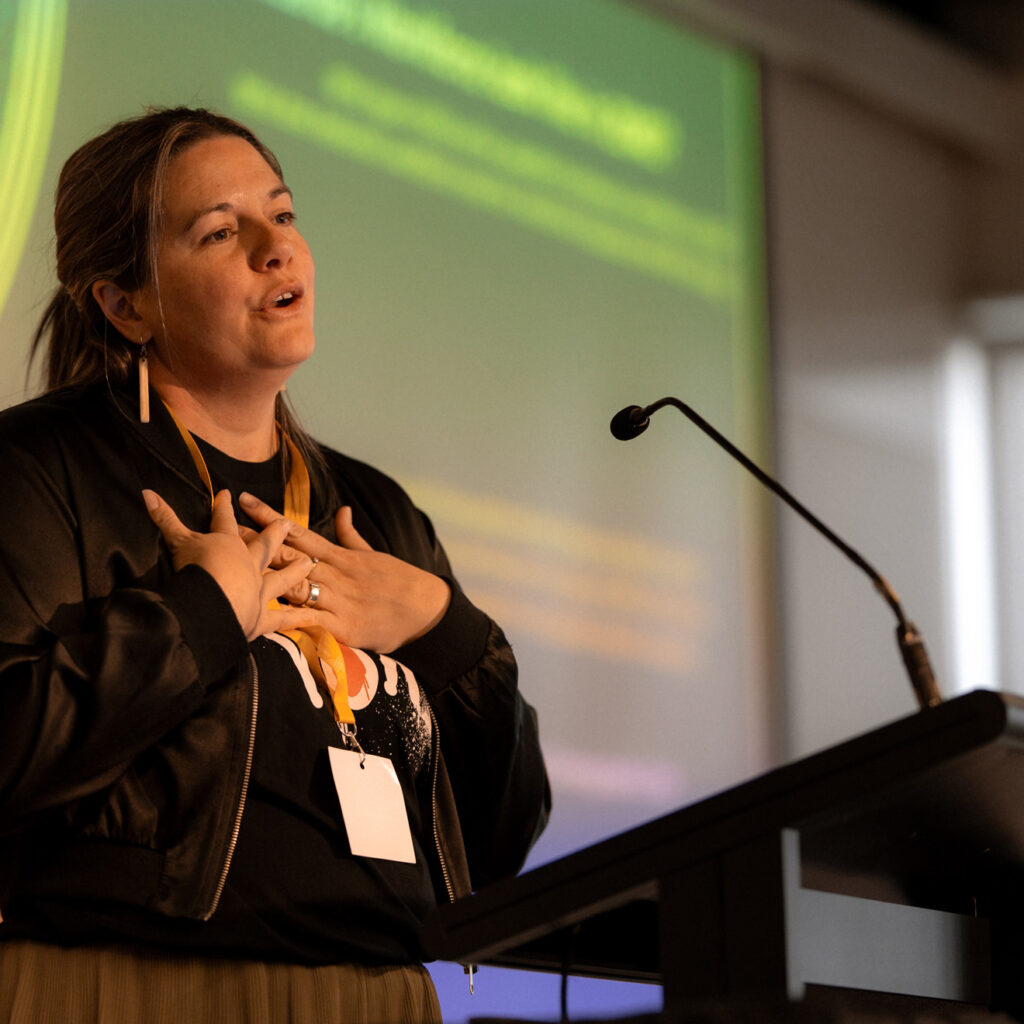 A woman with light brown hair, wearing a black jacket and a gold lanyard, speaks passionately at a podium. Her hands are held near her chest, and she has a serious but heartfelt expression. Another person, partially visible, stands behind her. A green slide with yellow text is projected on the screen in the background.