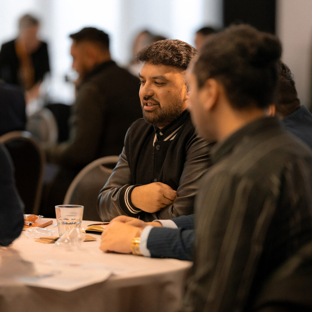 A group of people sit at a table in conversation. In the center, a man with short black hair and a beard, wearing a black varsity-style jacket, speaks while leaning forward slightly. Two others at the table listen intently, one with his back to the camera. The table has water glasses and notebooks scattered on it.