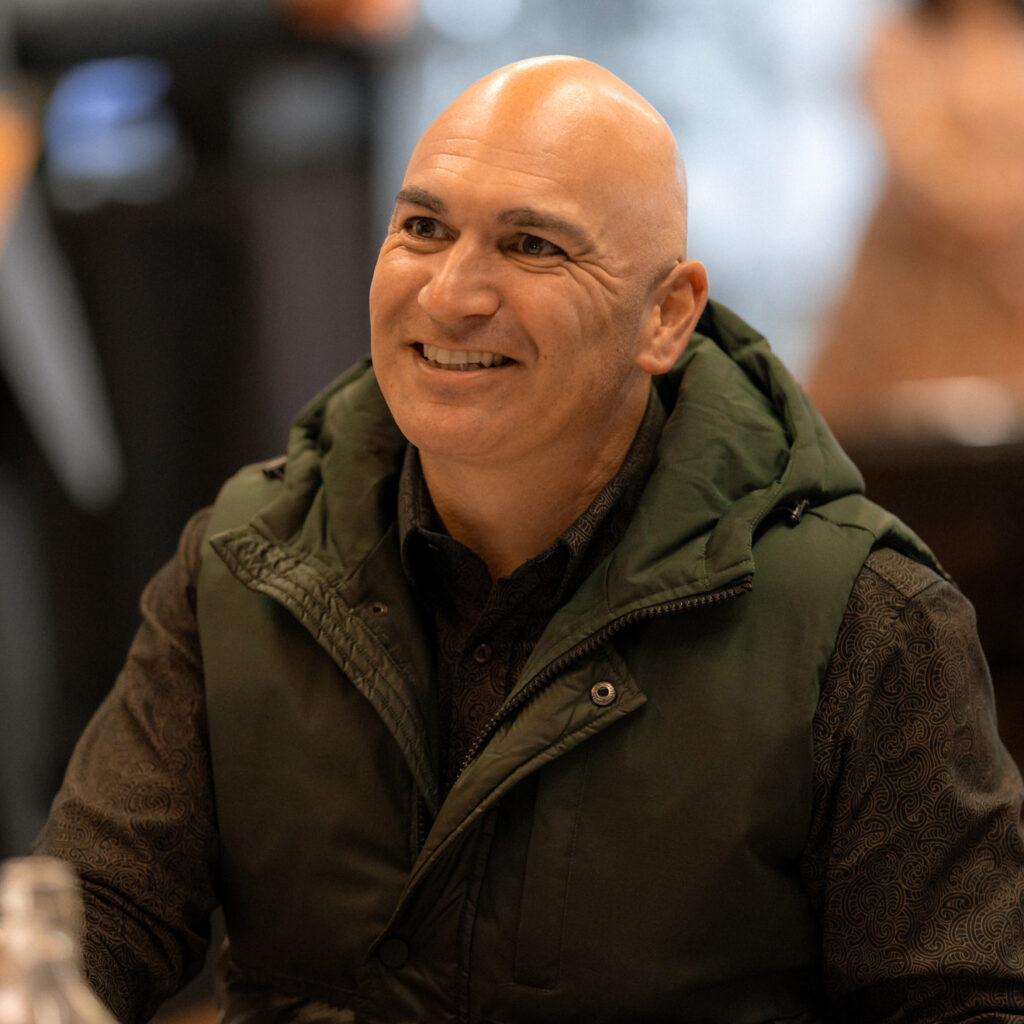 A smiling bald man in a dark green vest sits at a table, looking engaged in conversation. He has a warm expression, and the background shows a blurred setting of an event with people and tables.