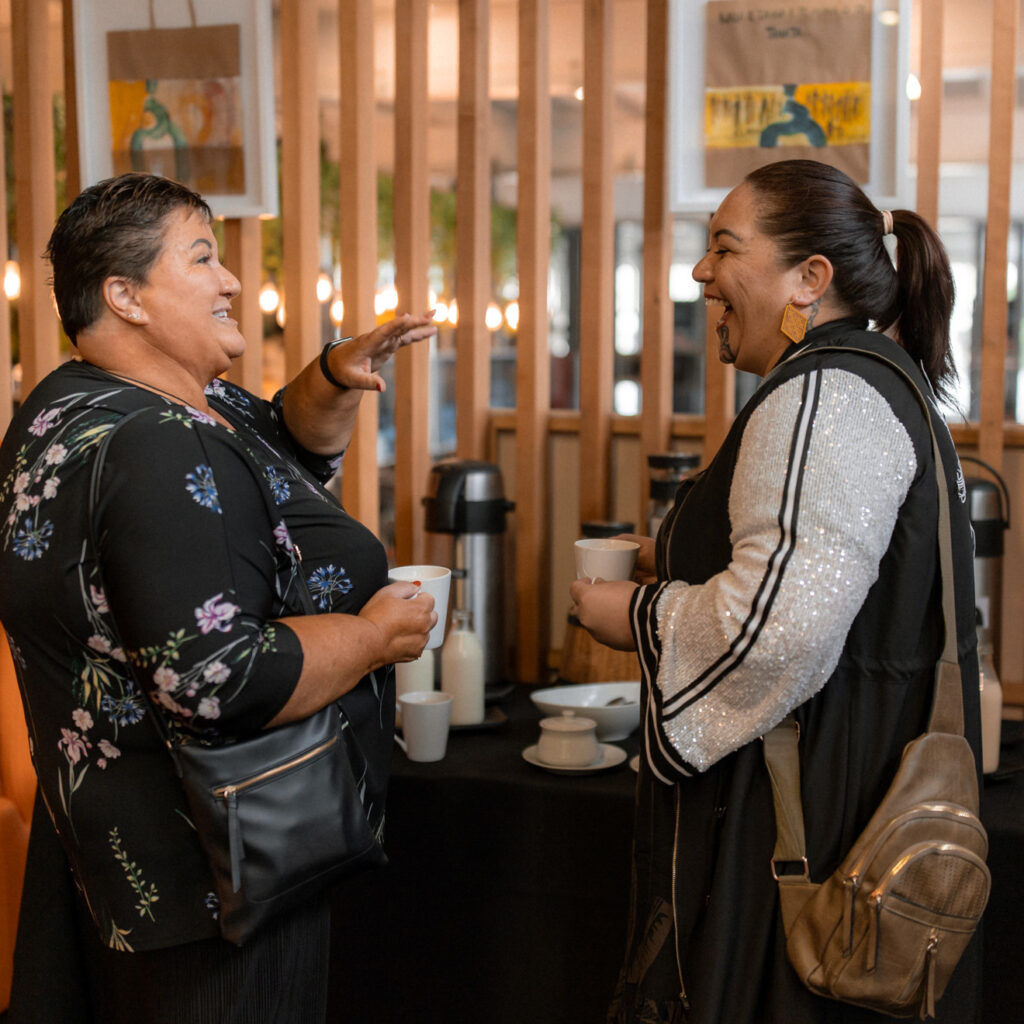 Two women holding coffee cups, laughing and chatting near a beverage station.