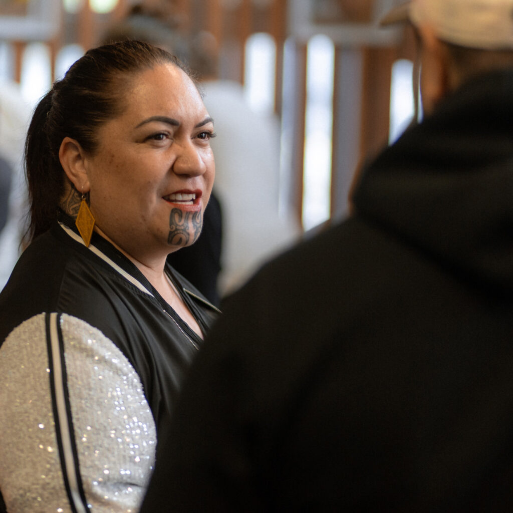 A woman with traditional moko kauae tattoo smiling and talking to someone off-camera.