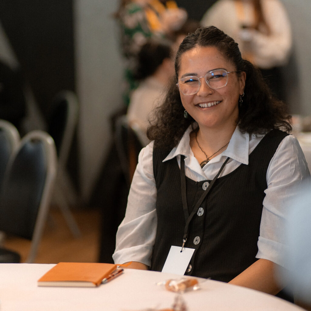 A seated woman with glasses smiling warmly at the camera during a gathering.