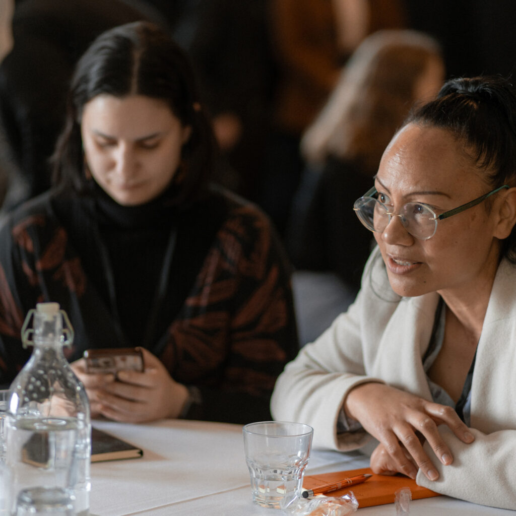Two women at a table, one looking at a phone while the other actively listens and engages.