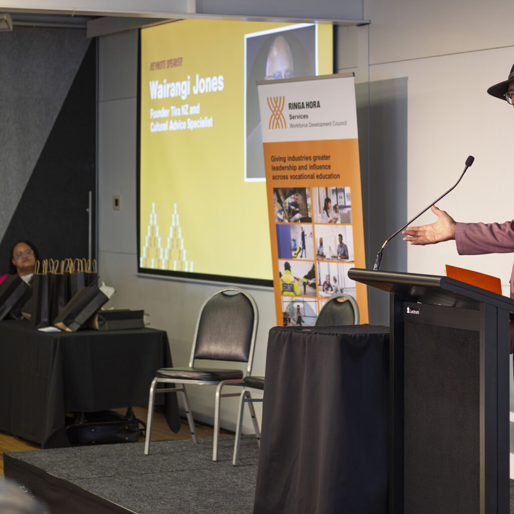 A man in a pink blazer and black hat is giving a speech at a podium with a microphone. Behind him is a large screen showing the text "Wairangi Jones," his title, and a yellow background. Two people are sitting at a table to the left, one smiling. A banner for Ringa Hora Workforce Development Council is visible.